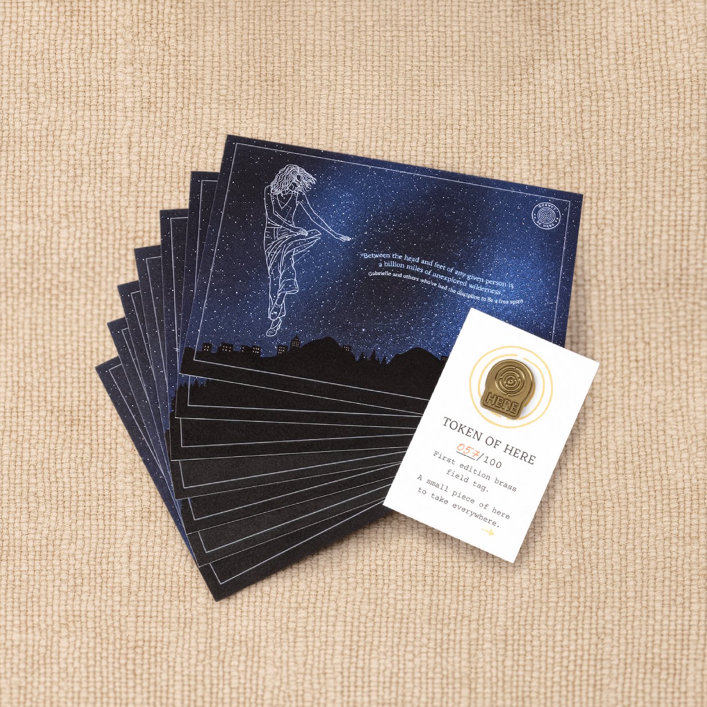 Product photo of a brass pin badge placed on top of night-sky star artwork from the Kit of Here.