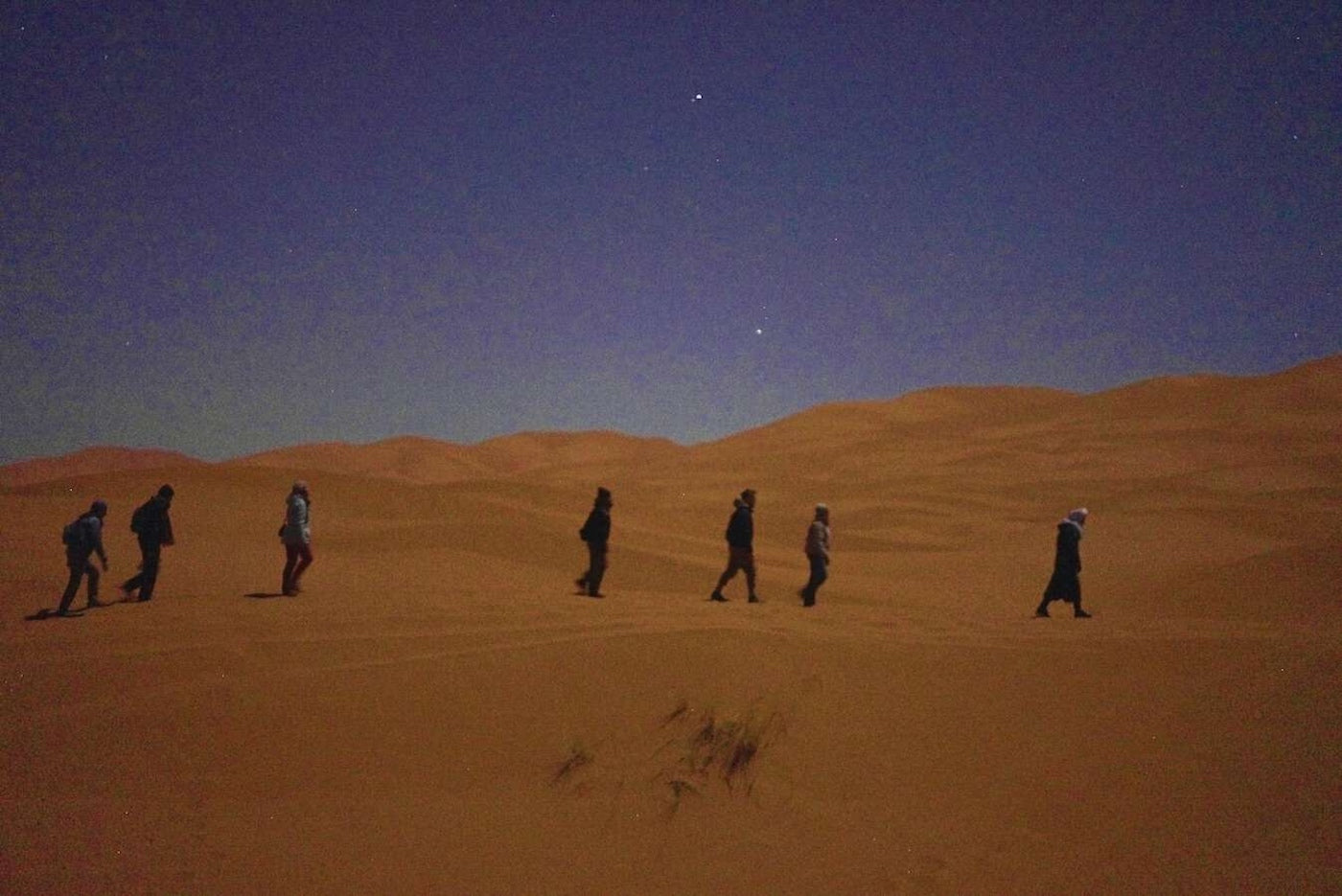 Group of people walking across Sahara desert sand dunes at night under stars, symbolising journey and belonging.